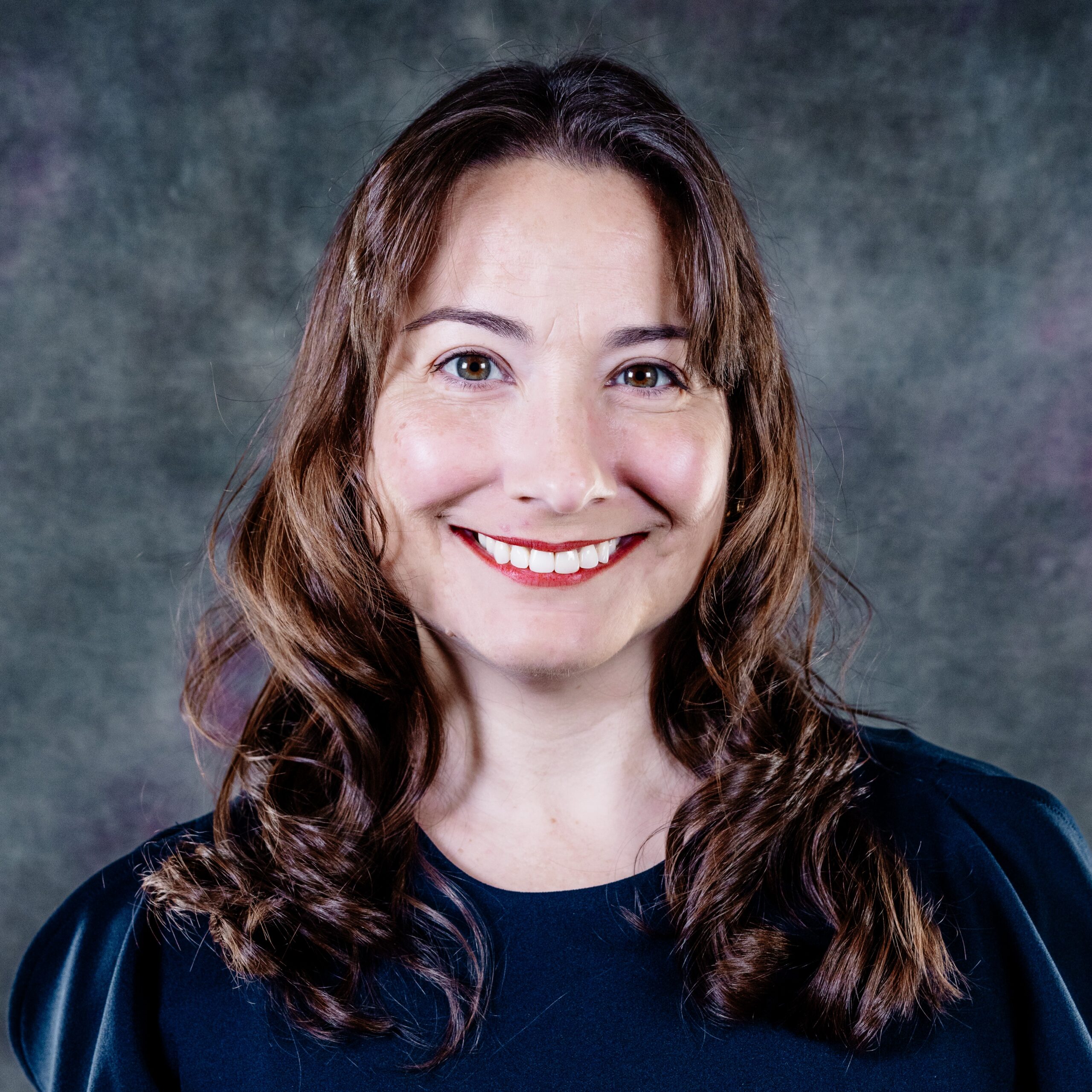 Woman smiling with dark brown hair, red lipstick, and a blue blouse.