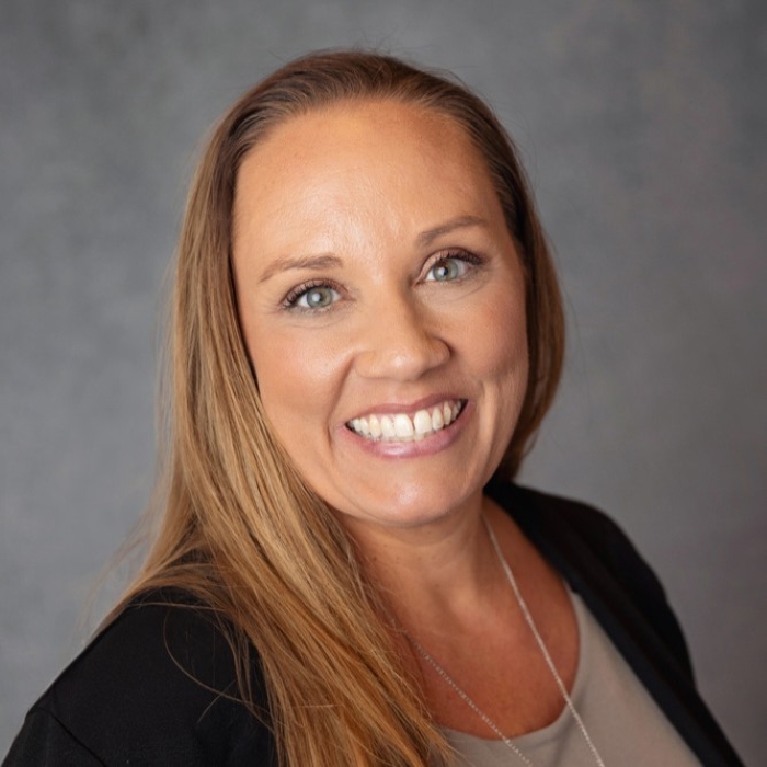 Woman smiling with blue eyes, straight brown hair and gray background.