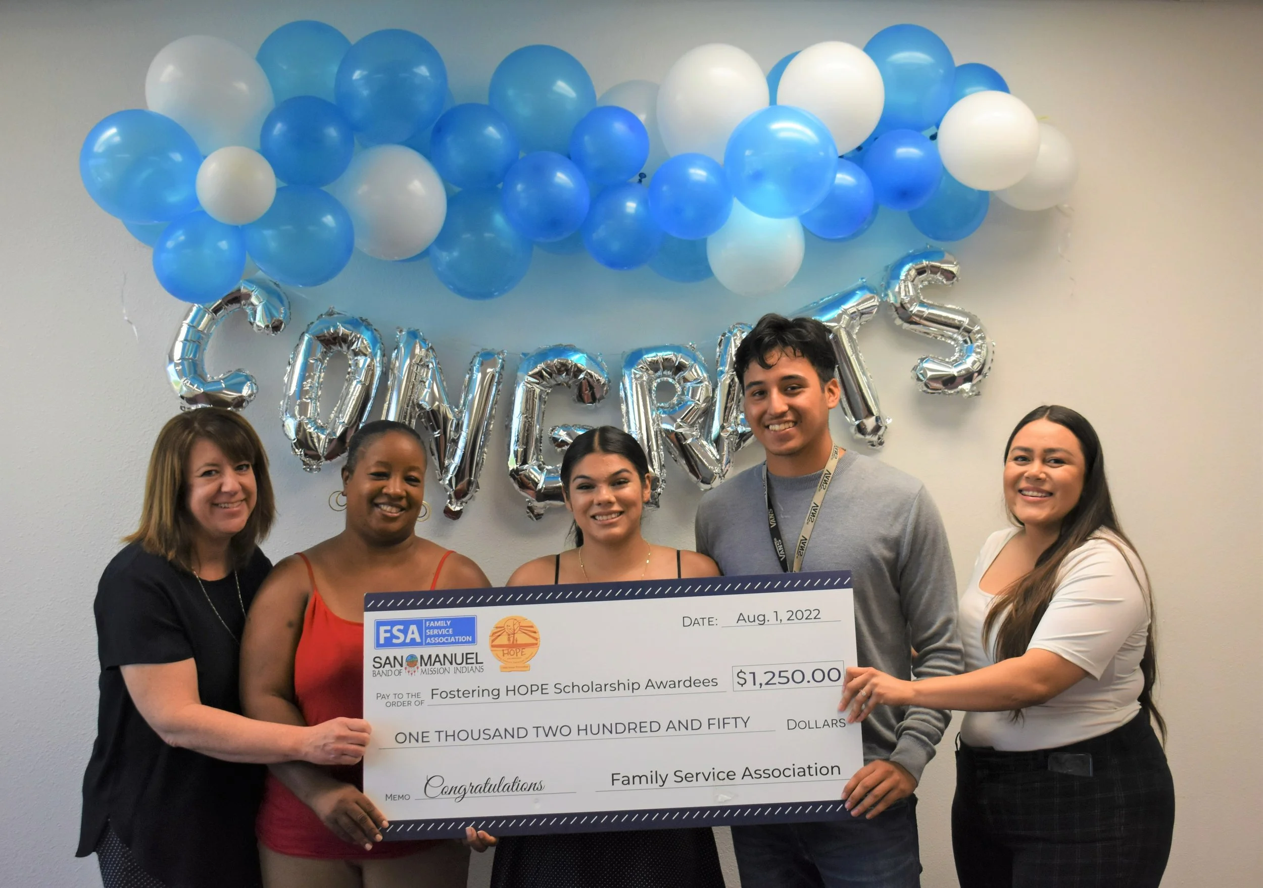 五个人面带微笑,手里拿着一张价值 1,250 美元的大额奖学金支票,支票后面是写有“CONGRATS”的气球字母。