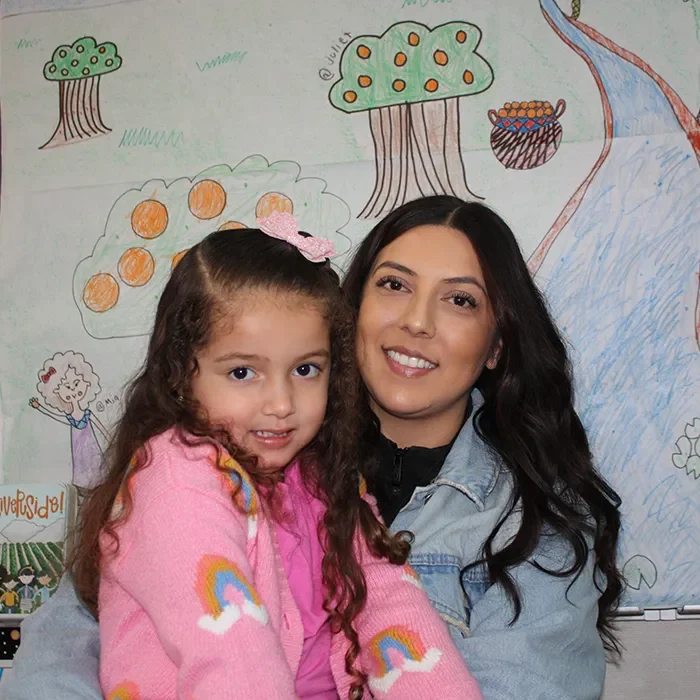 A woman and young girl smiling in front of a colorful, child-drawn mural with trees and rainbows.