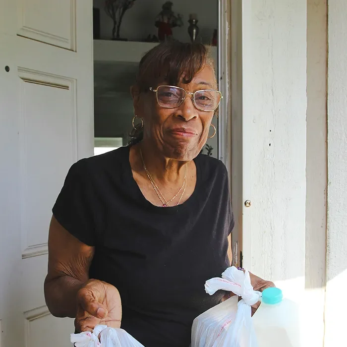 Older woman in glasses and a black shirt stands in a doorway, holding grocery bags and a jug of milk.