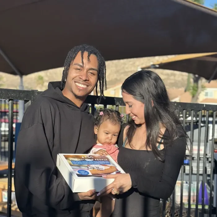 A smiling couple holds a baby and a box of cookies outdoors on a sunny day.