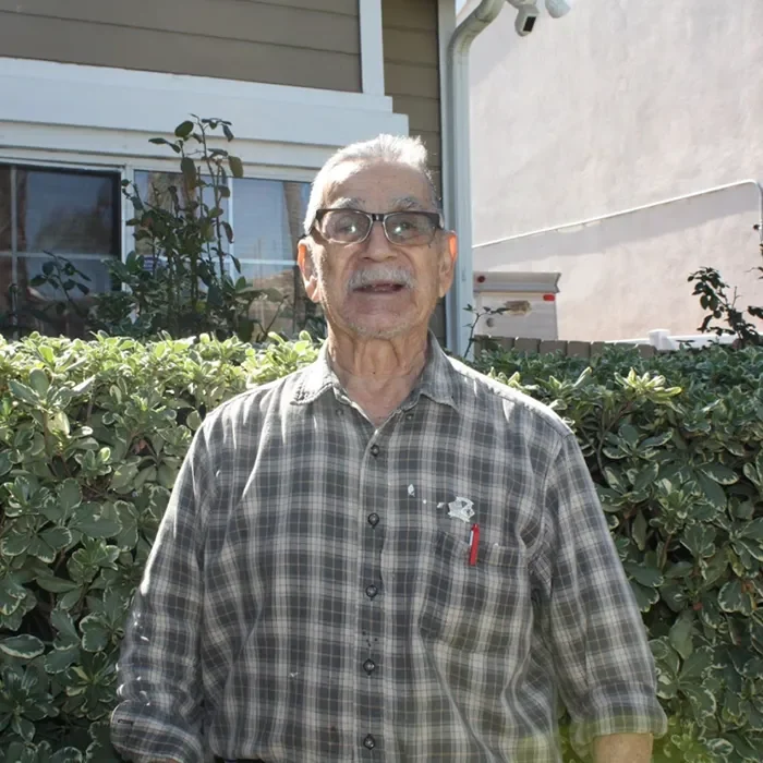 Elderly man in plaid shirt standing and smiling in front of green bushes and a house.