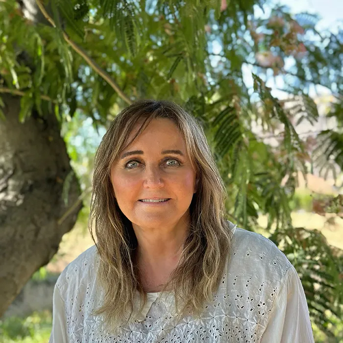A woman with long light brown hair smiles outdoors under a leafy tree, wearing a white embroidered blouse.