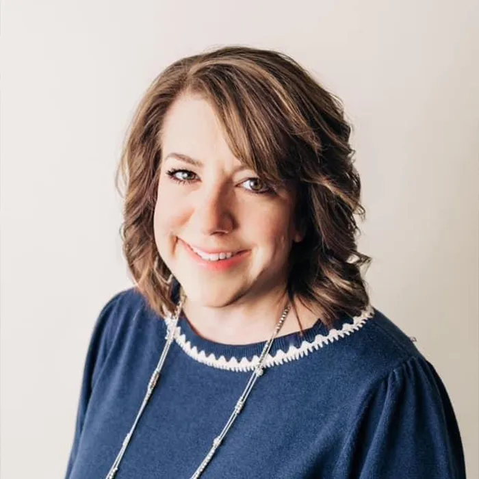 Woman with wavy brown hair in a blue top smiling in front of a plain light background.