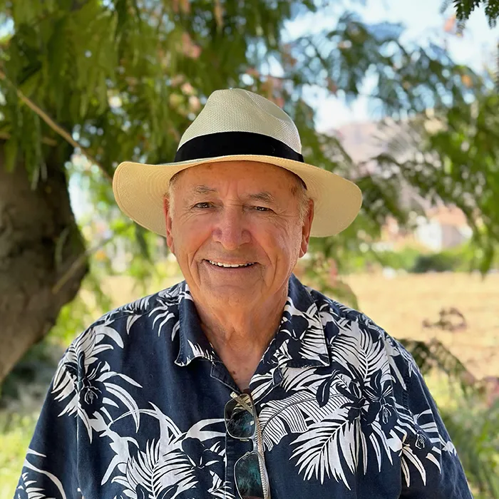 Smiling older man in a straw hat and floral shirt stands outdoors with trees and sunlight in the background.
