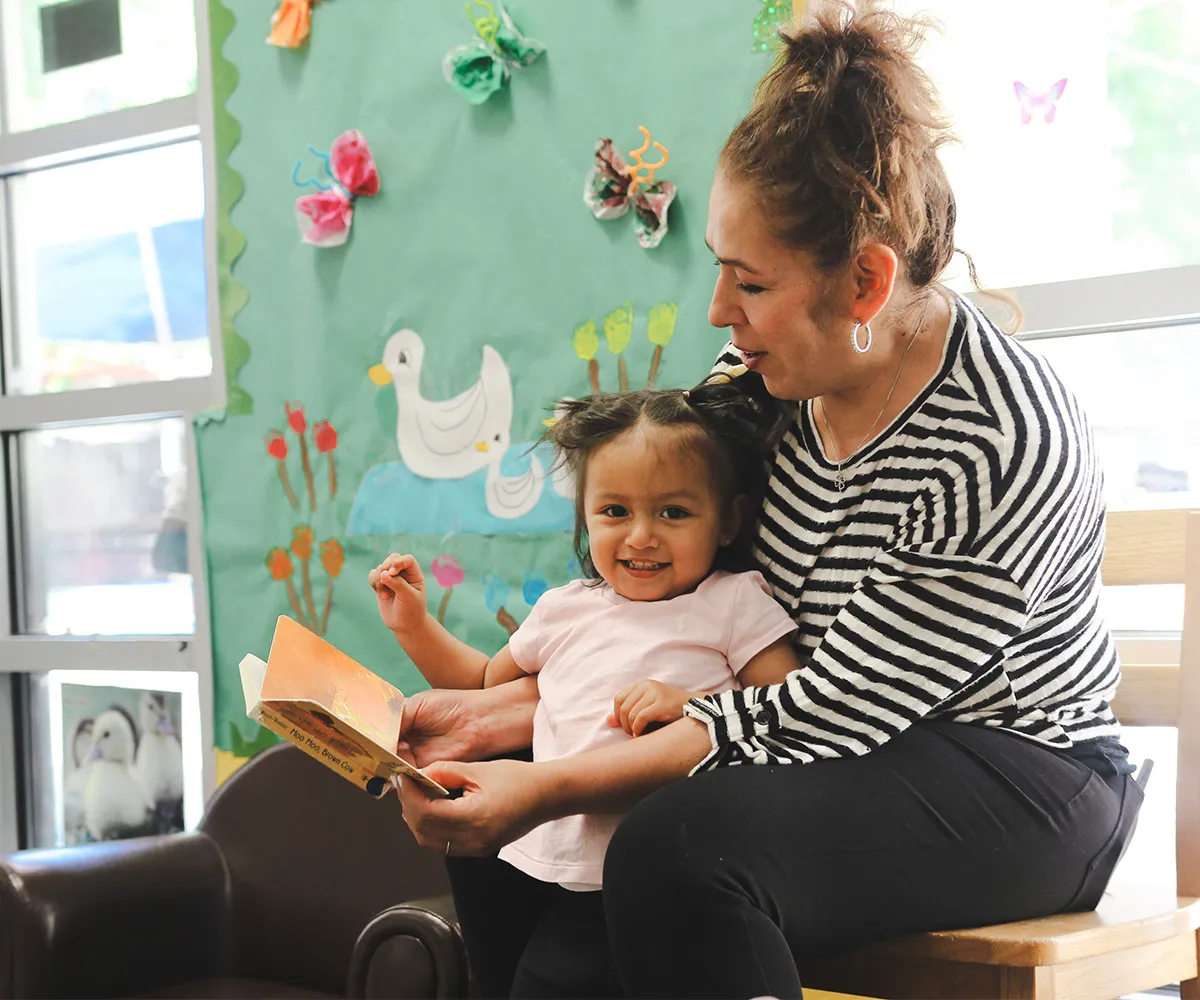 Adult reading a book to a smiling toddler in a colorful classroom decorated with paper art and drawings.