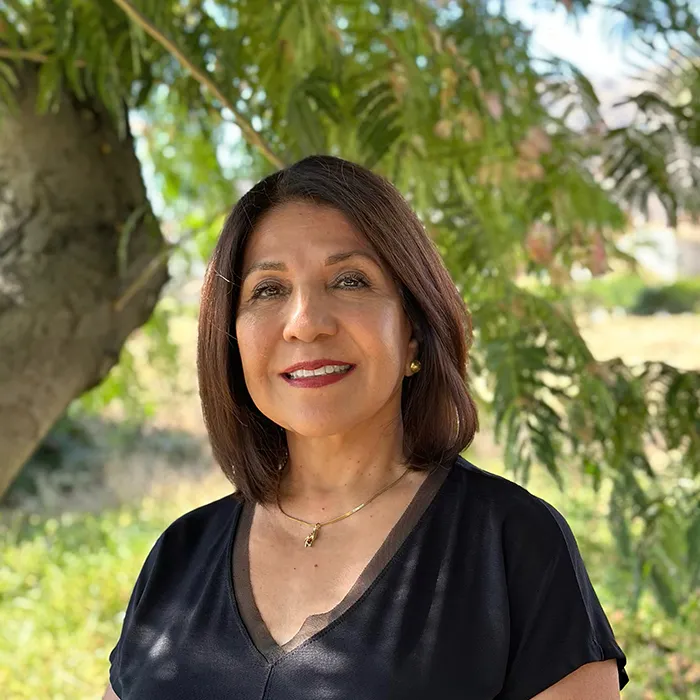 Woman with shoulder-length brown hair, wearing a black top, smiling outdoors in front of a leafy tree.