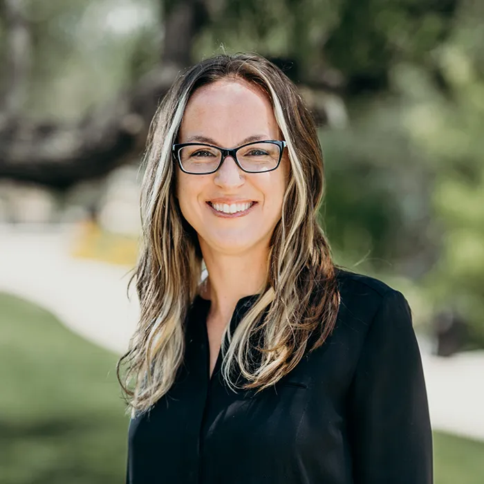 Woman with long wavy hair and glasses, smiling outdoors in a park setting with trees in the background.