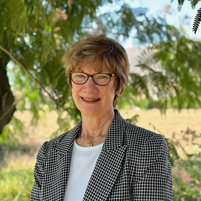 A woman with short hair and glasses, wearing a checkered jacket, smiles outdoors with trees in the background.