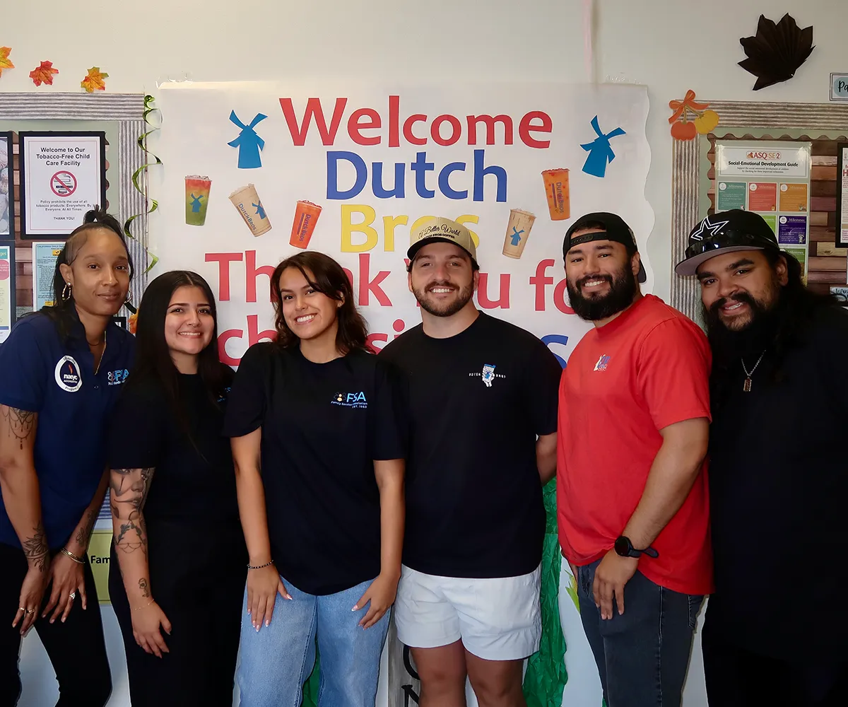 Six people smile in front of a colorful "Welcome Dutch Bros" sign on a wall decorated with artwork.