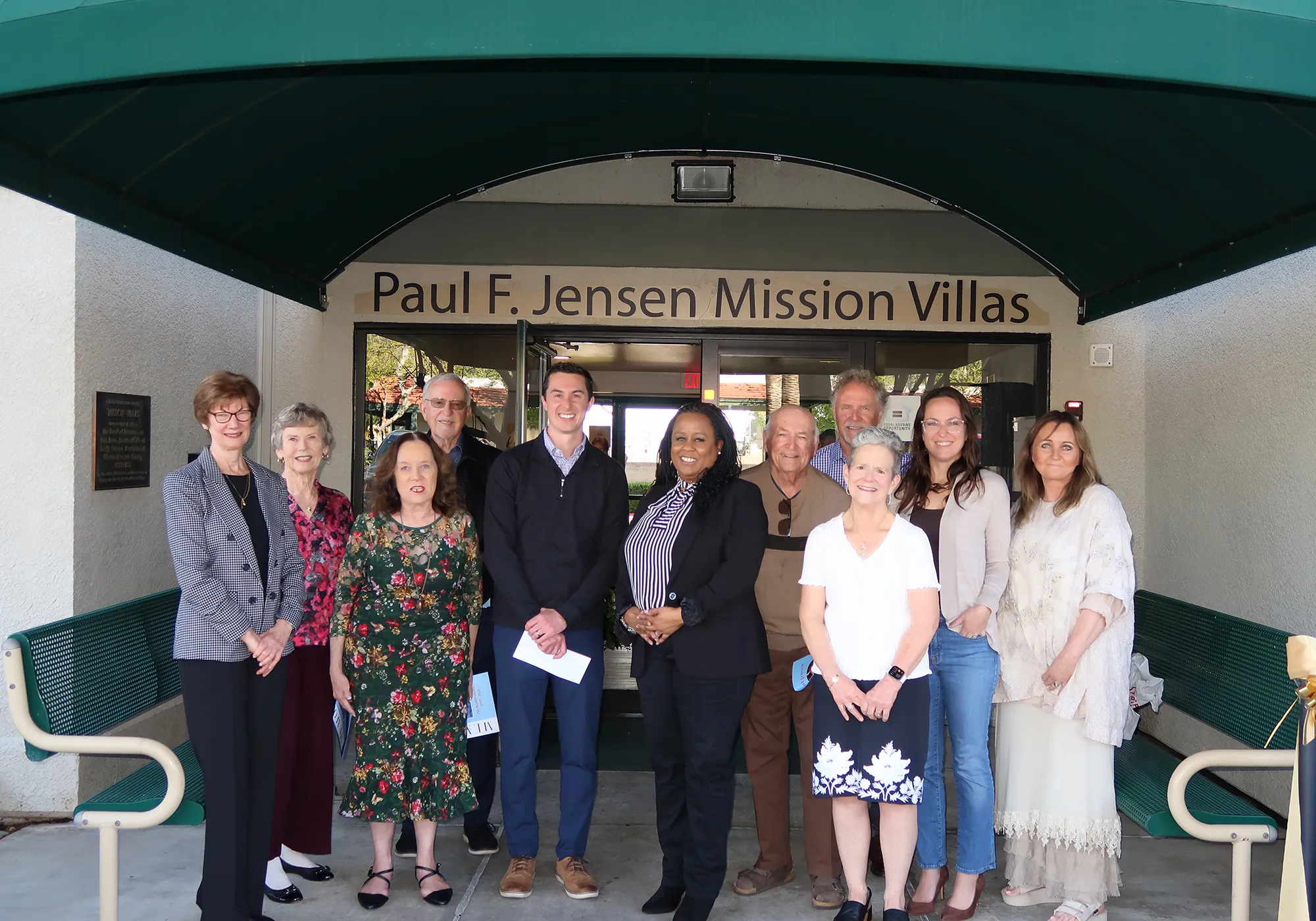 Twelve people stand smiling in front of the Paul F. Jensen Mission Villas building entrance.