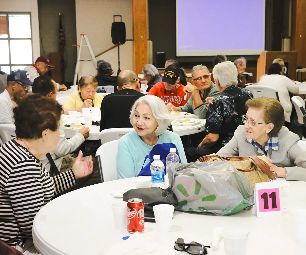 A group of seniors sit at tables, eating and chatting in a community center or event hall.