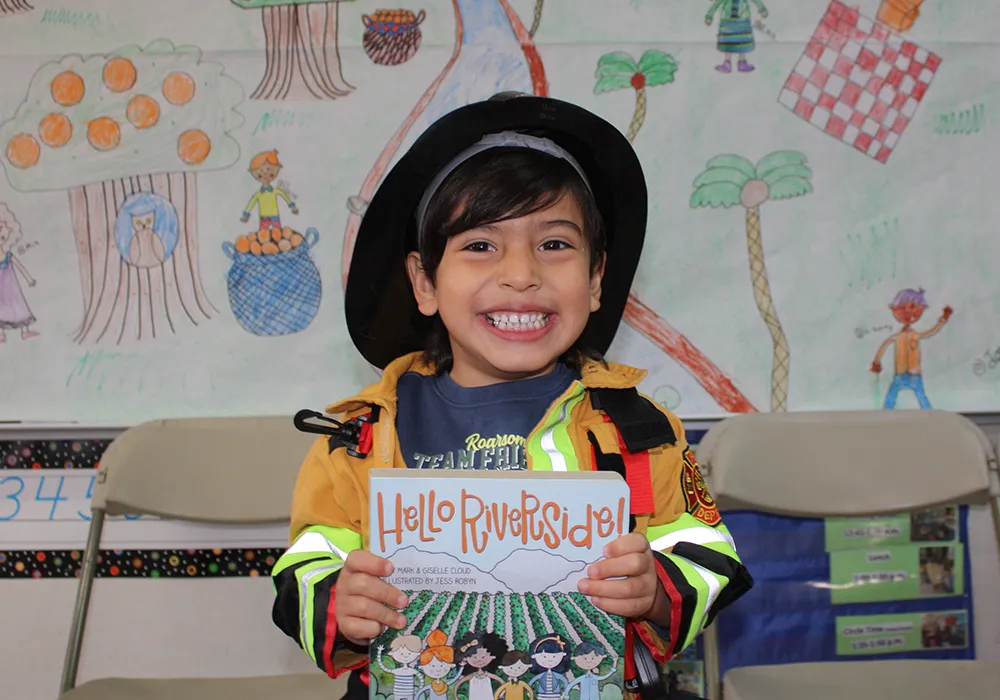 Smiling child in a firefighter costume holds a “Hello Riverside!” book in front of a colorful drawing.