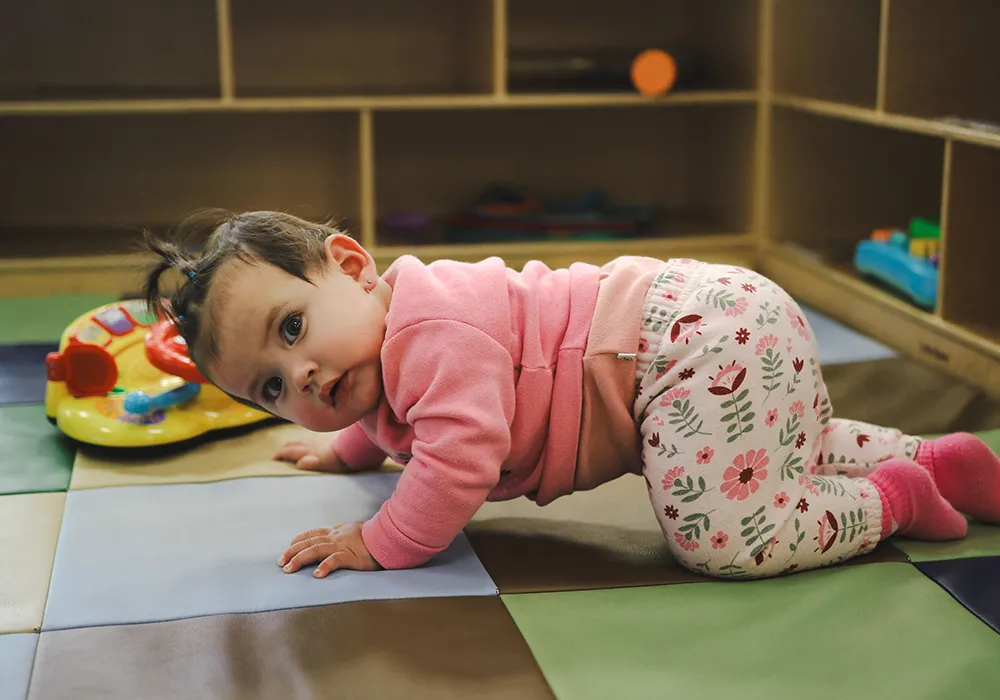 A baby in pink clothes crawls on a play mat, looking toward the camera, with toys nearby.