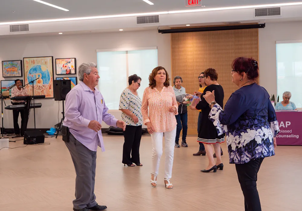 A group of older adults dance together in a brightly lit community center with art on the walls.