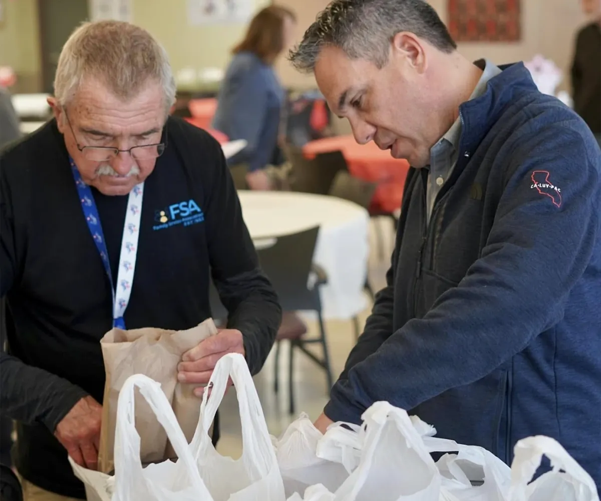 Two men pack food items into white plastic bags at a table covered with bags indoors.