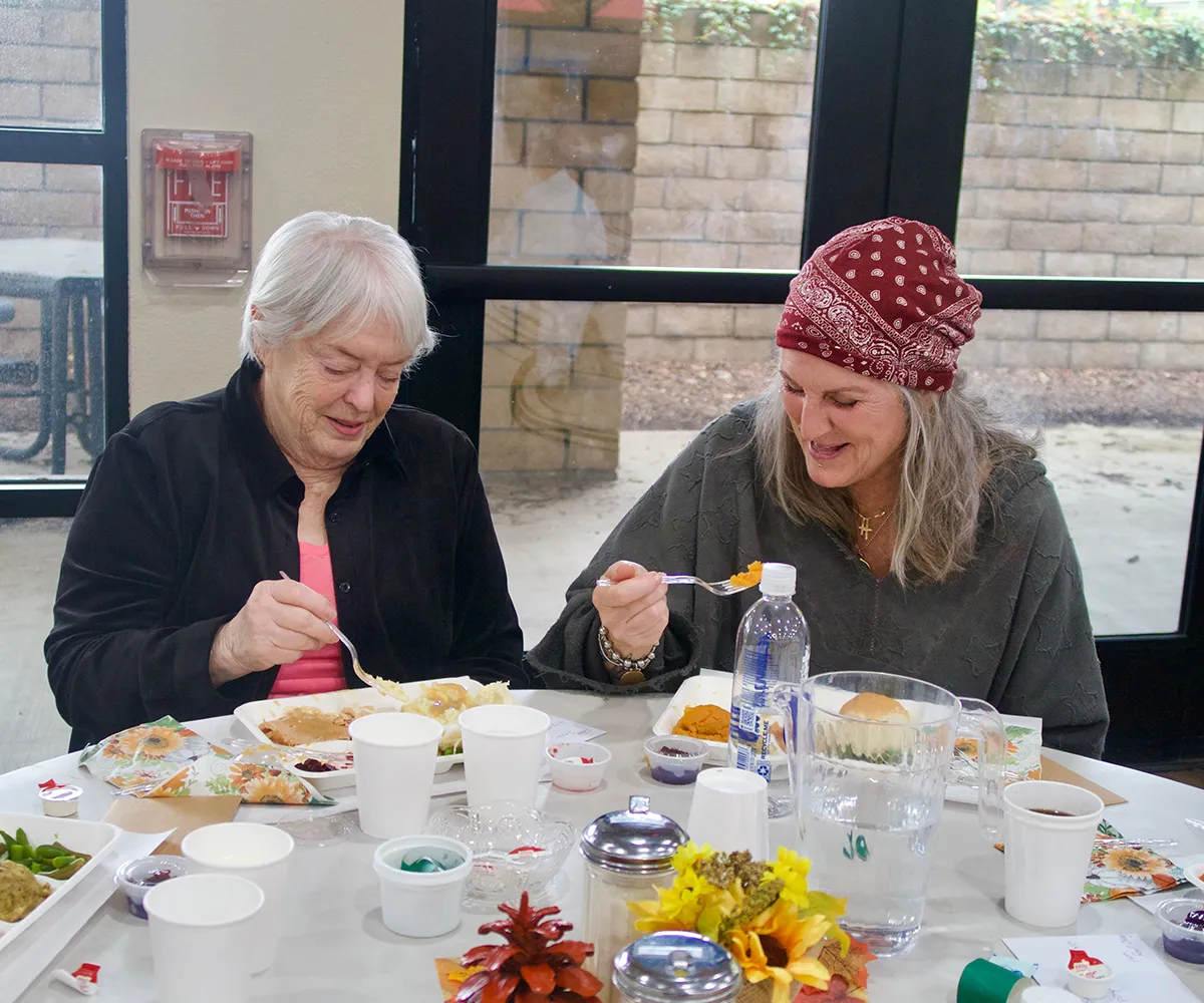 Two women sit at a table, smiling and eating a meal together, with food and drinks in front of them.