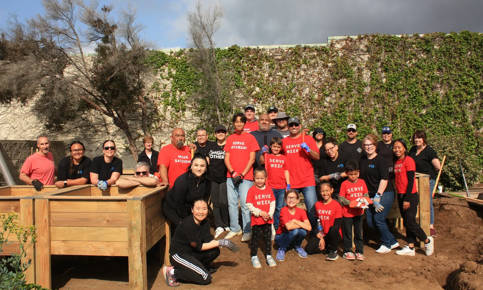 A group of people pose together outside by a raised garden bed, some wearing matching "Serve Week" shirts.
