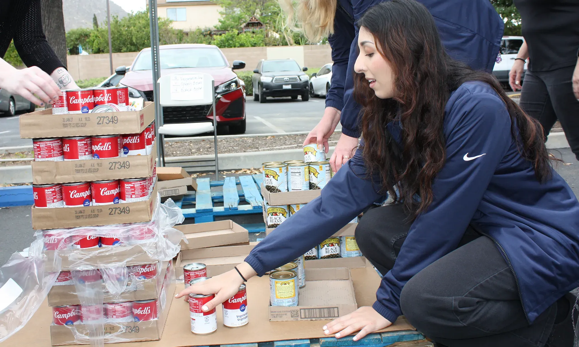 Woman crouches, organizing cans of soup on a pallet outdoors with others helping in the background.