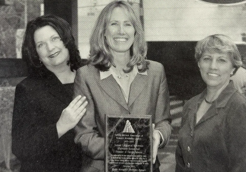 Three women smiling; the center woman holds a plaque. They are dressed in business attire.