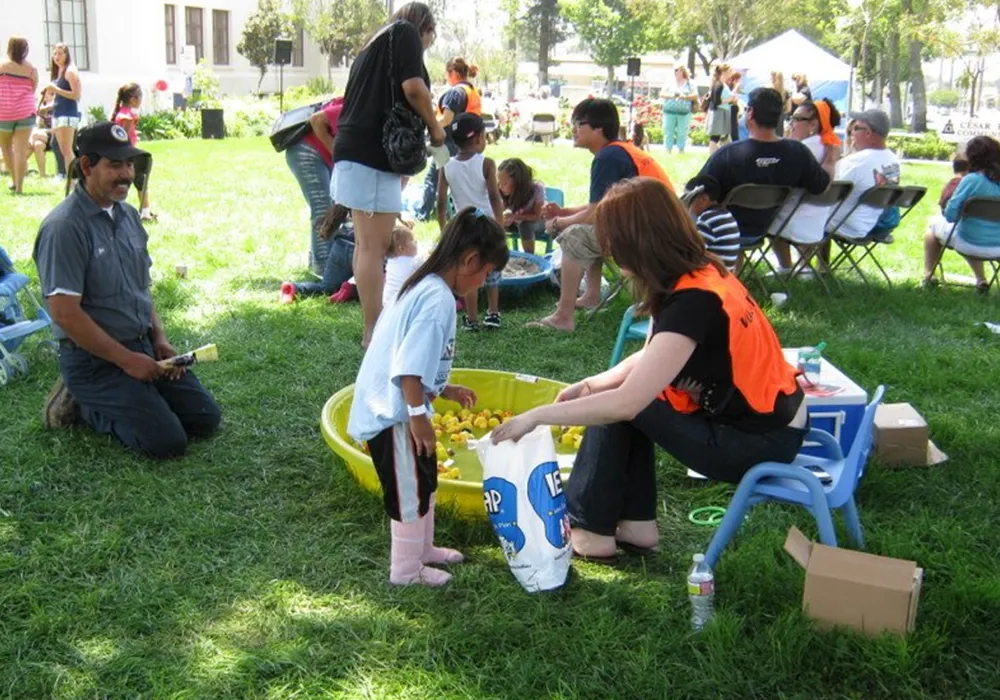 A woman helps a young girl at an outdoor event with a duck pond game, while others gather around.