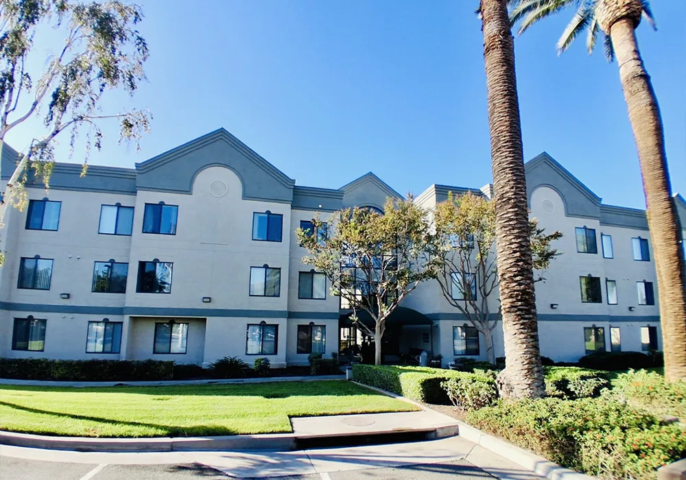Three-story beige building with tall palm trees, green lawn, and clear blue sky in the background.