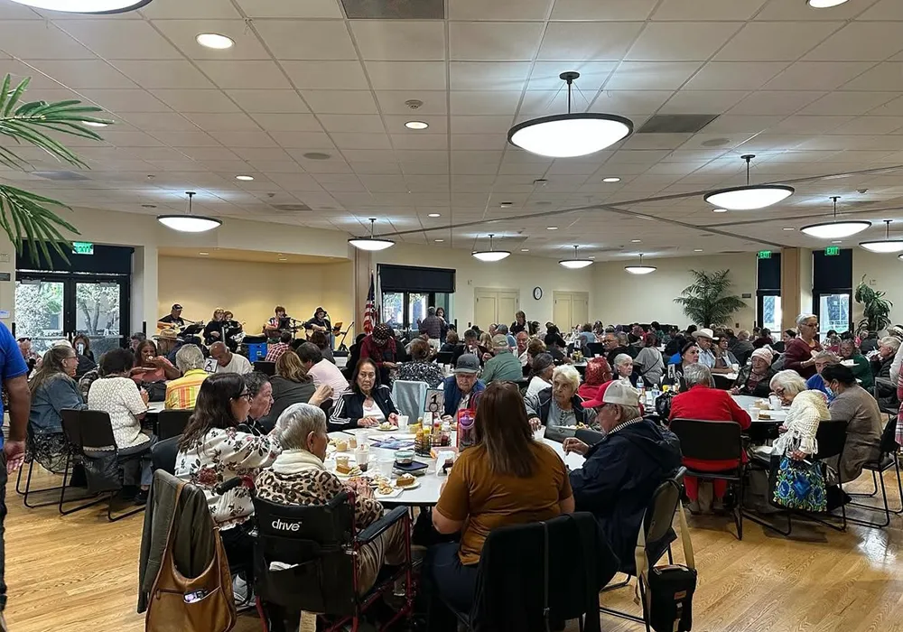 Large group of people sitting at tables in a brightly lit community center, sharing a meal together.