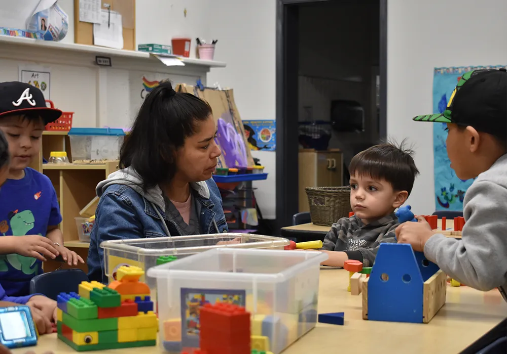 Woman and three children playing with colorful building blocks at a table in a classroom.