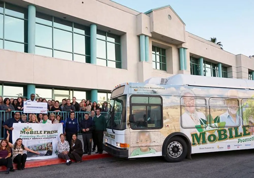 A group poses by a bus with “Mobile Fresh” signage outside a modern building in daylight.