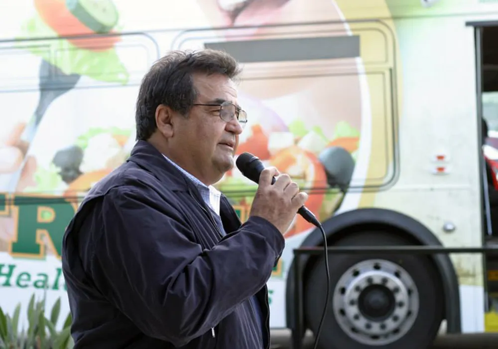Man speaking into a microphone outdoors, with a bus featuring food images in the background.