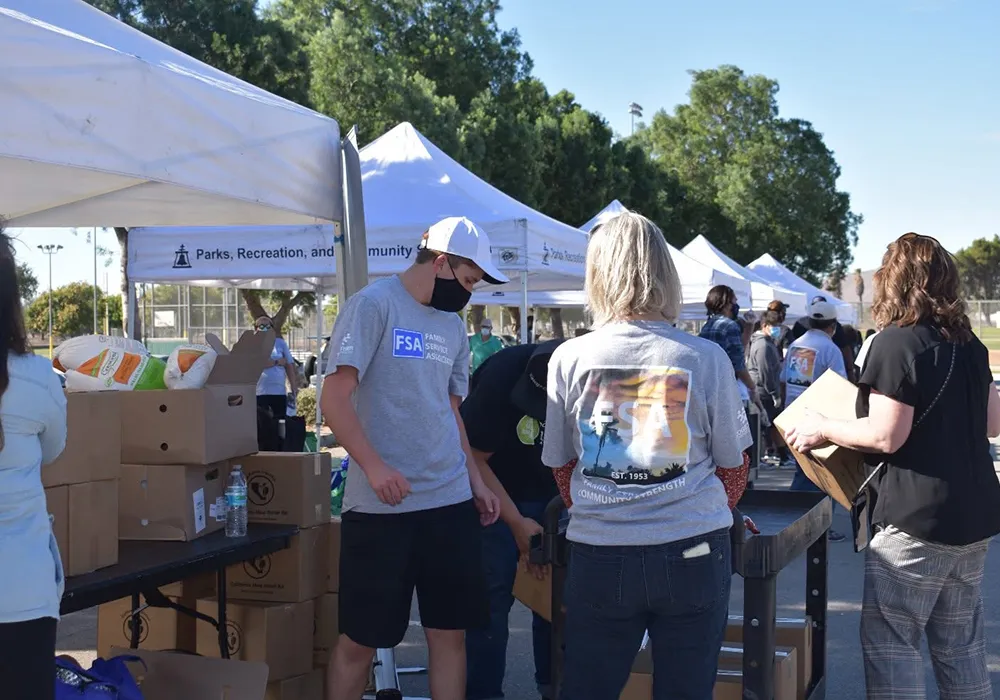 People sorting boxes at an outdoor community event under white tents on a sunny day.