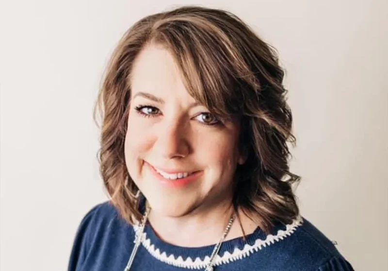 Smiling woman with wavy brown hair wearing a navy blue top and necklace, posing against a light background.