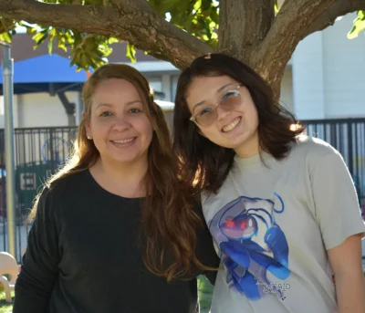 Two people smiling and standing together outside under a tree on a sunny day.